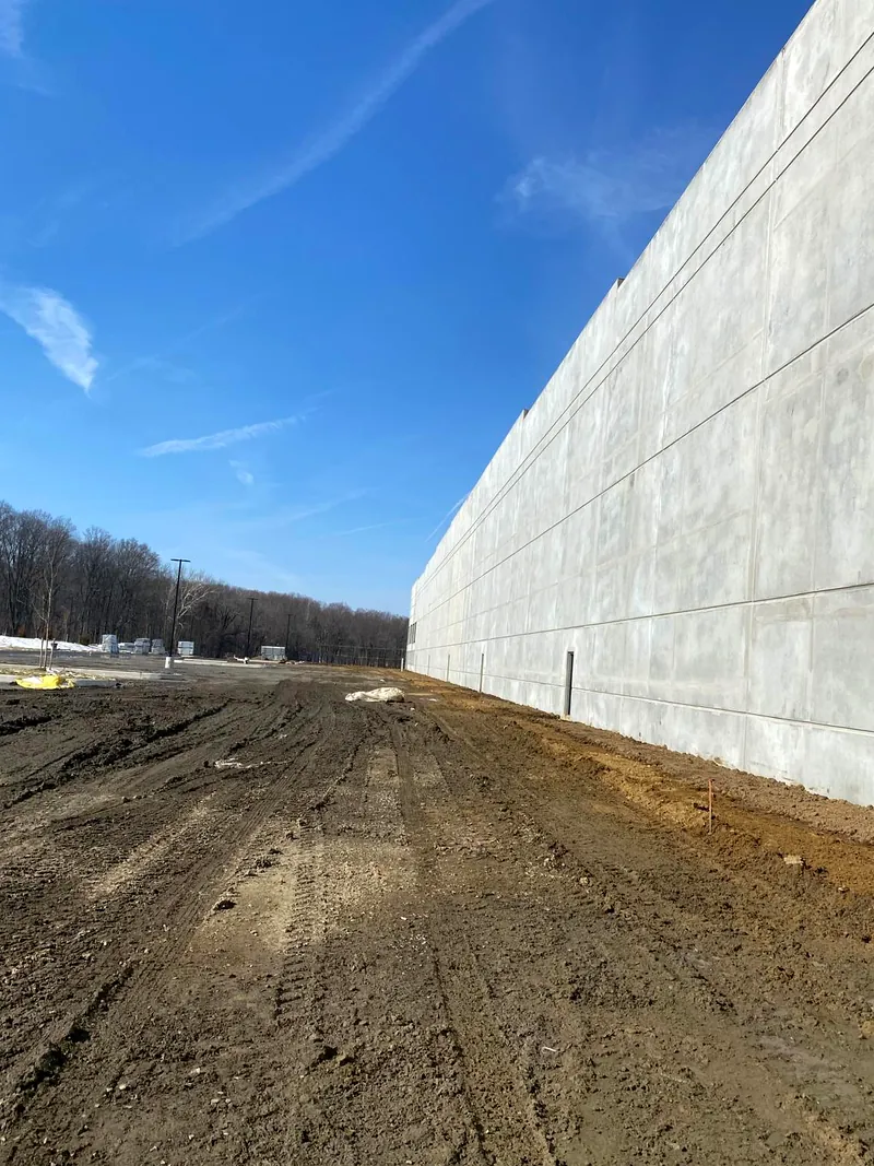 Long run of bare grey tilt-up concrete panels beside a graded dirt lot under a blue sky
