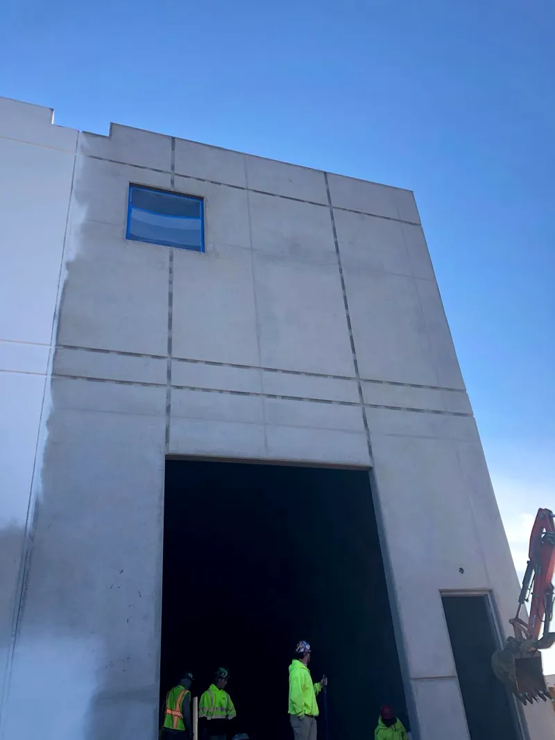 Tall bare concrete panels at a building entry with an open doorway, workers in high visibility vests and an excavator on site
