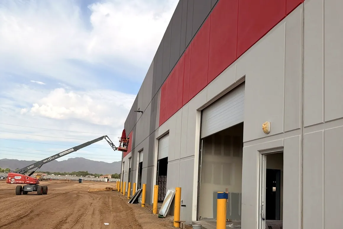 Tilt-up commercial facility exterior with a painted red accent band, grey panels, dock doors and yellow bollards