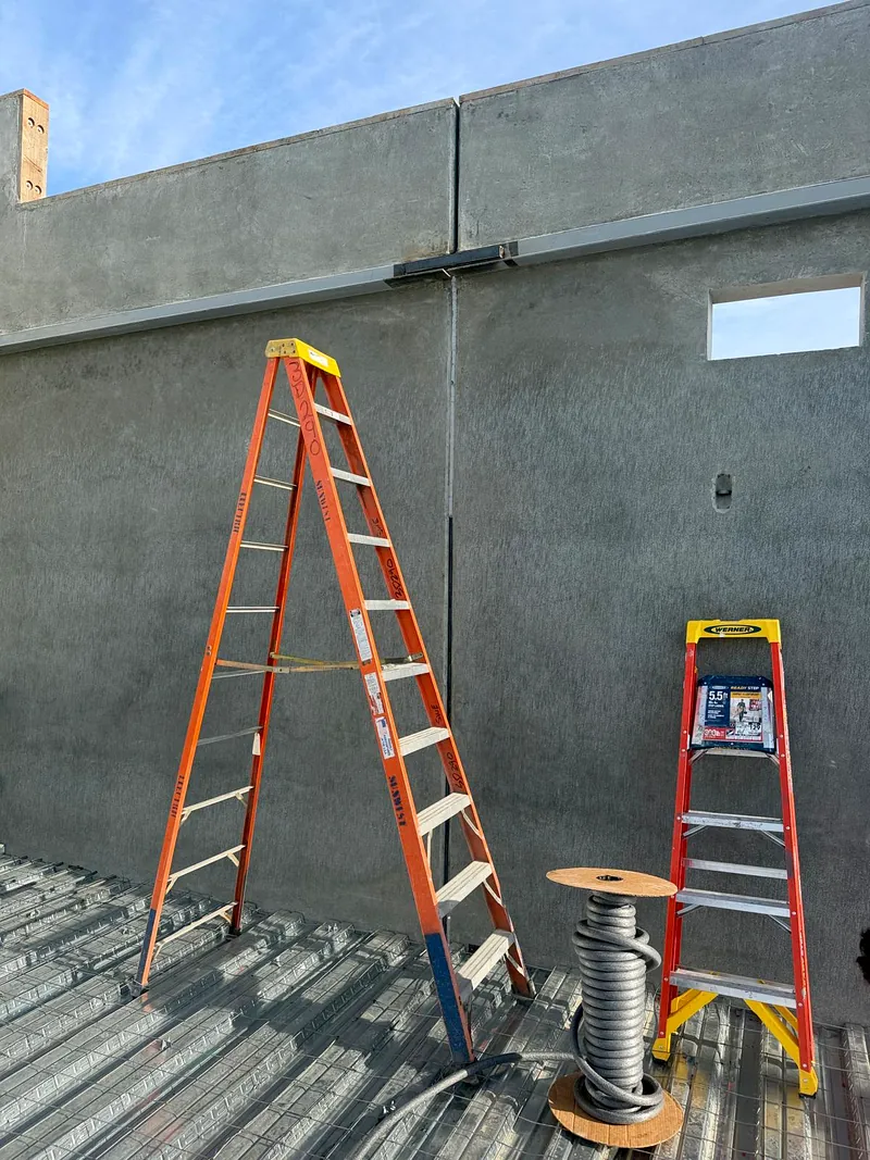 Bare grey parapet wall at roof deck level with two ladders, a cable spool and metal roof decking