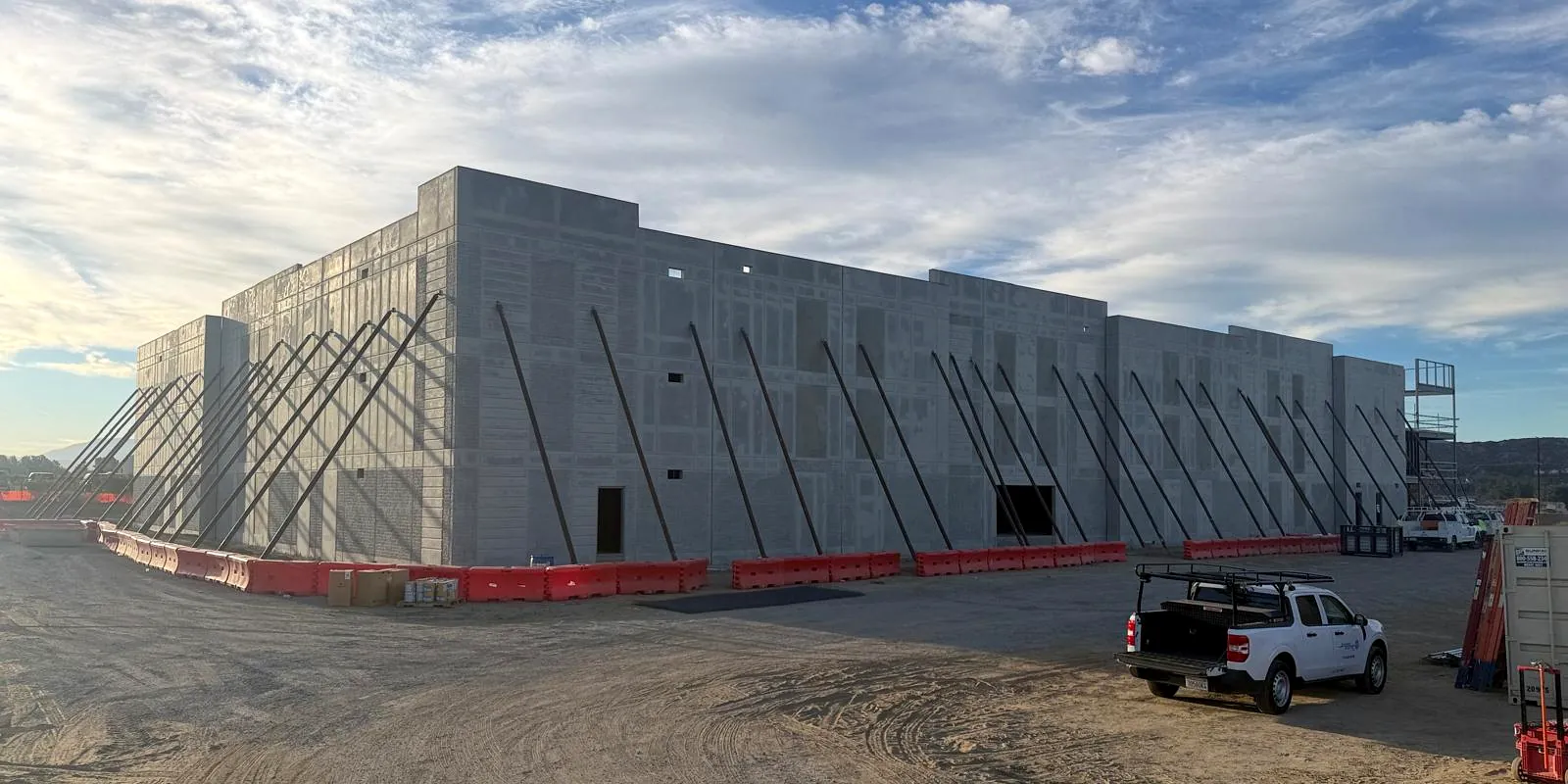 Wide view of a tilt-up concrete building shell with diagonal braces, all panels still bare grey concrete on a dirt lot